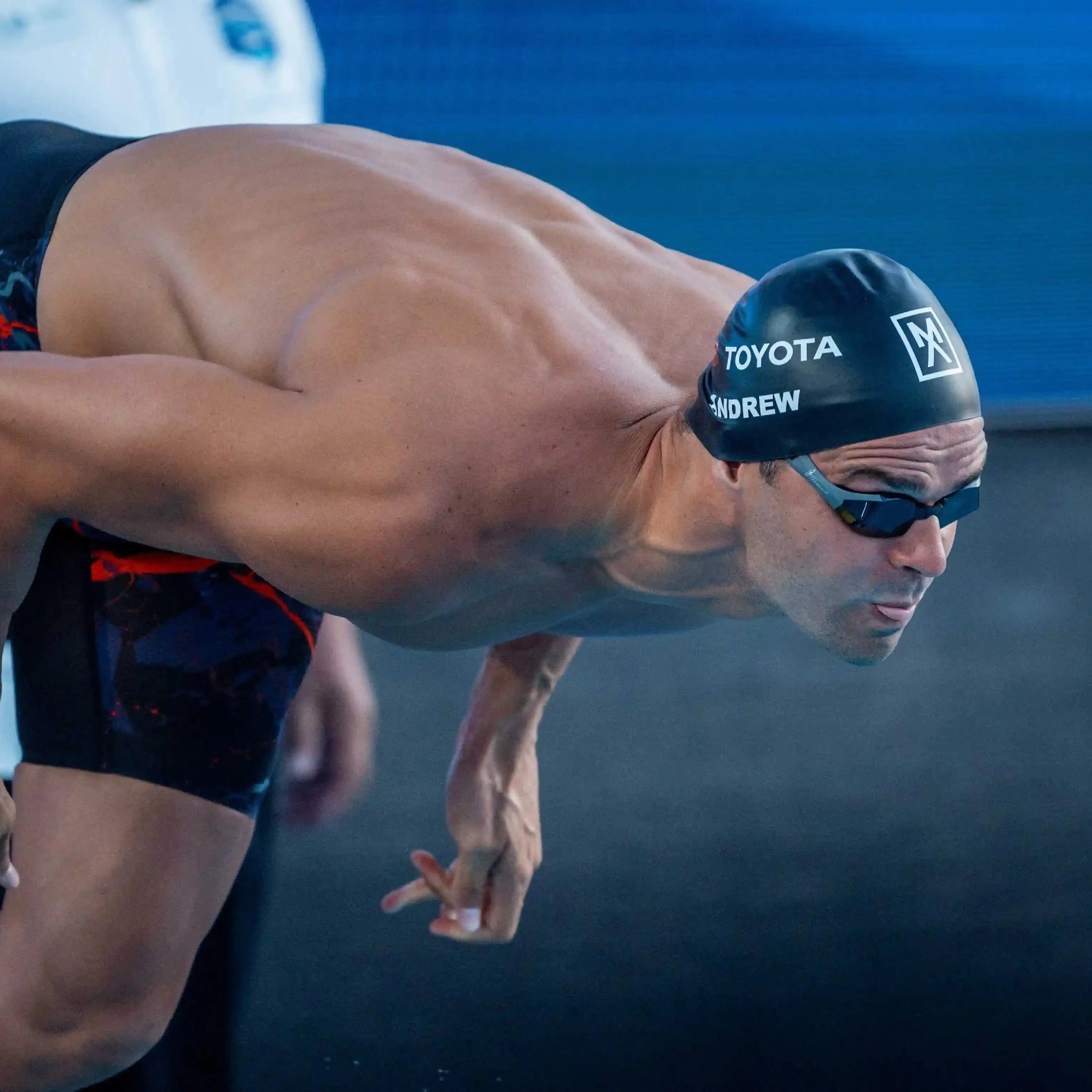 Michael Andrew in a competitive racing position, wearing a black swim cap with white 'ANDREW' branding, black goggles, and black swim trunks with red accents, captured mid-action against a dark blue pool background.