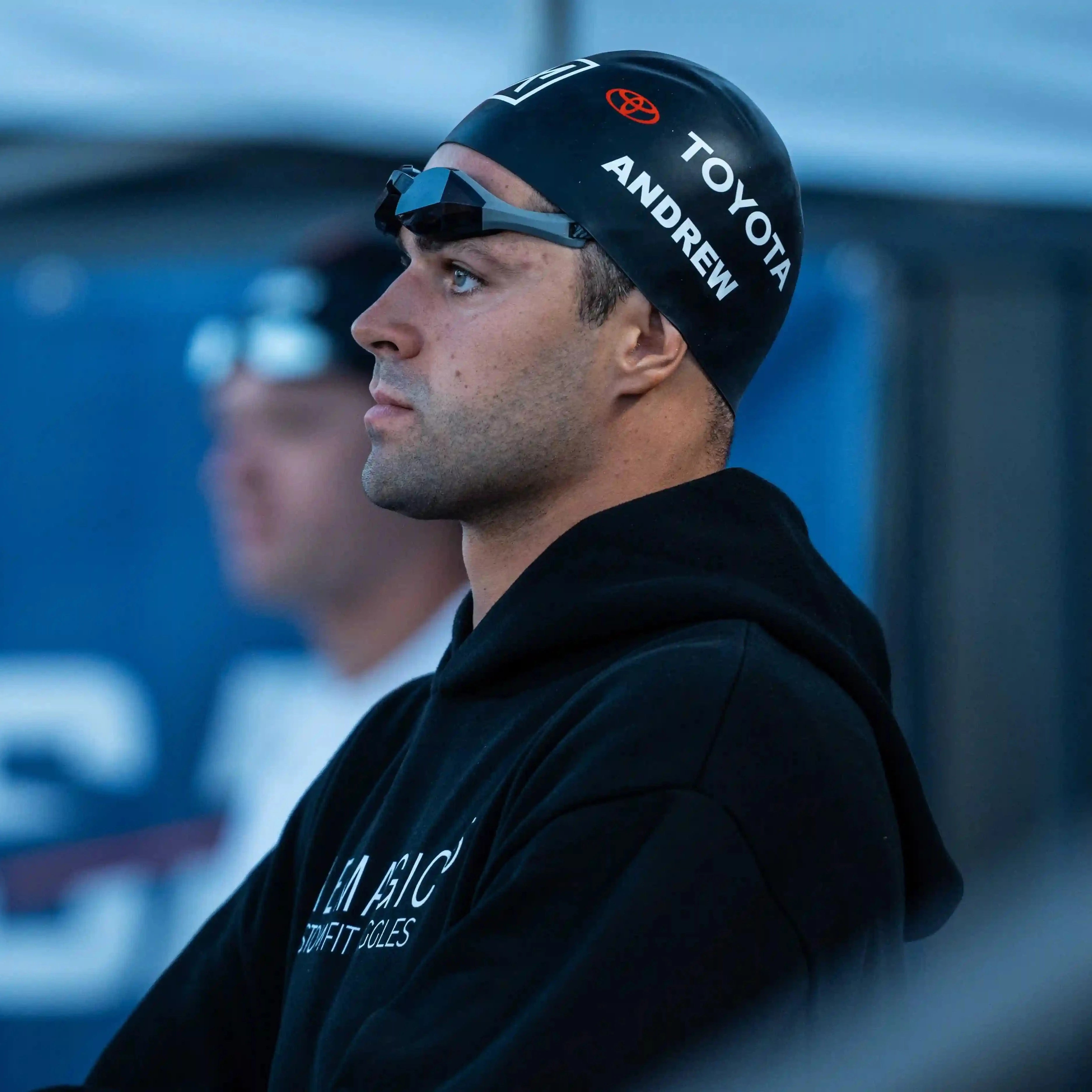 Michael Andrew a black hoodie and black swim cap, wearing THEMAGIC5 swim goggles, captured in a side profile at an outdoor event.