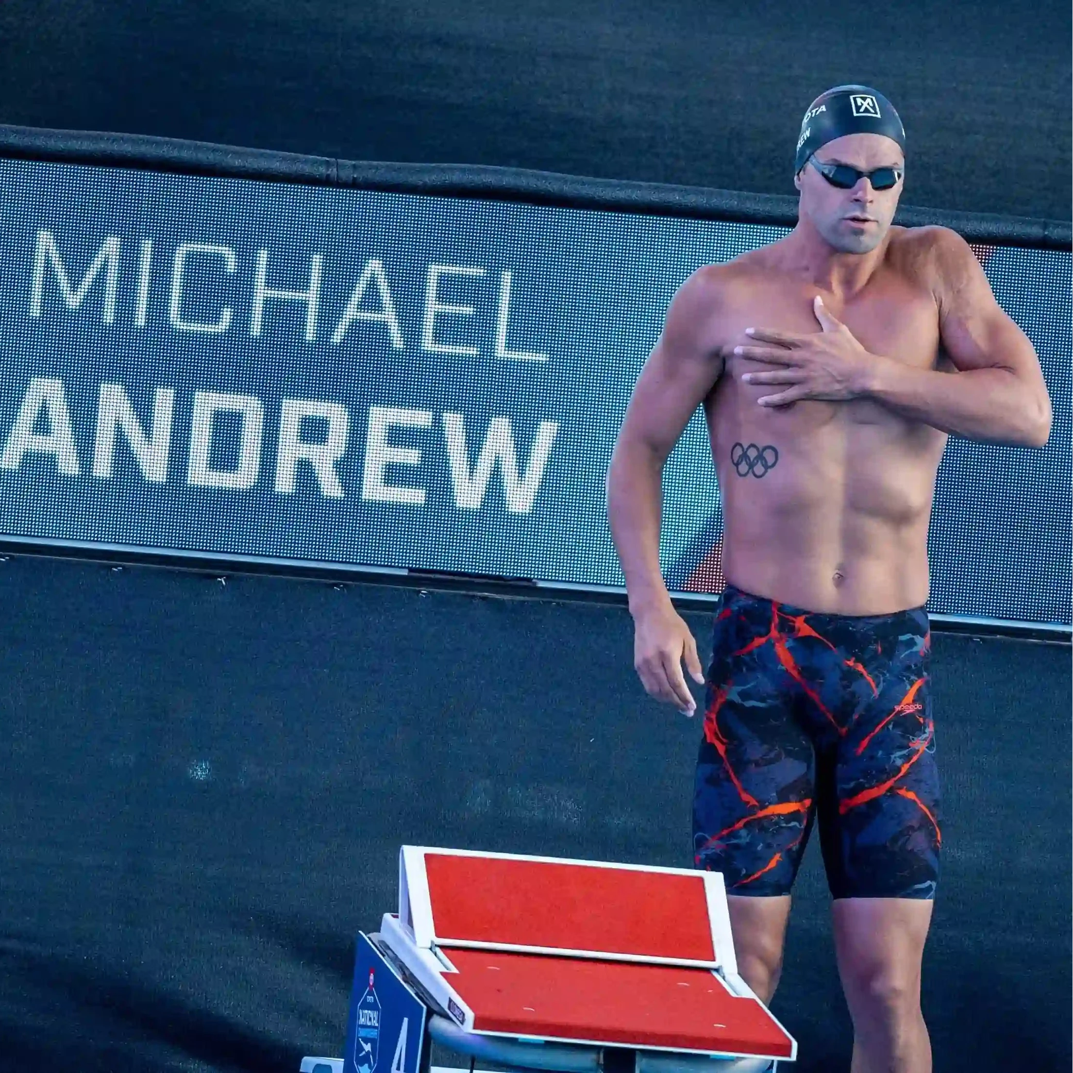 Michael Andrew, a professional swimmer, standing on a starting block at the pool. He is wearing a dark blue swim cap with a white logo, black THEMAGIC5 swim goggles, and dark blue patterned swim trunks with orange accents.